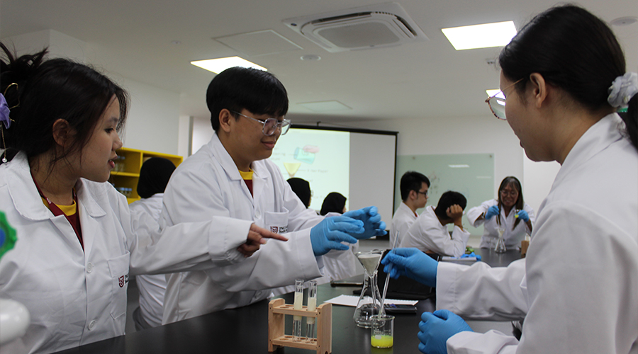 Nursing College Brunei | Male and female students from JPMC College of Health sciences, Brunei, in a classroom, learning to use and mix chemicals, female lecturer explaining, projector showing course slides, bench with test tubes and flasks.