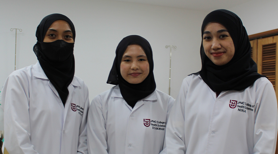Nursing College Brunei | Three female students, smiling, Asian, JPMC College of Health Sciences lab coat, black tudung black hijab, standing in classroom.
