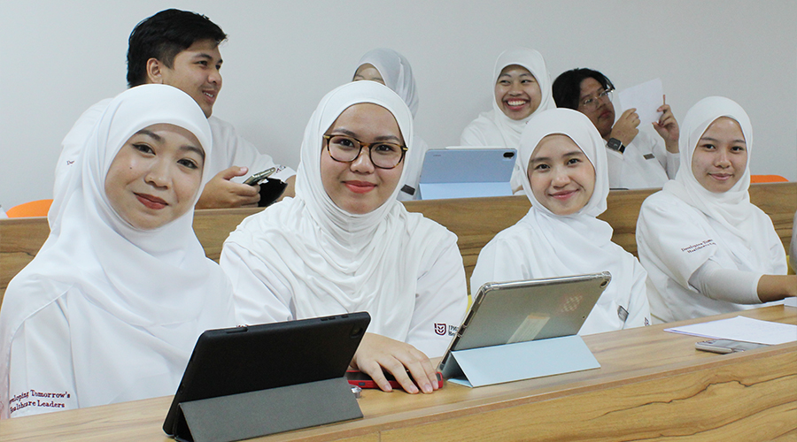 Nursing Course Brunei | Smiling female students male students glasses reading paper laughing using phones tablets studying nursing in classroom at JPMC College of Health Sciences Brunei