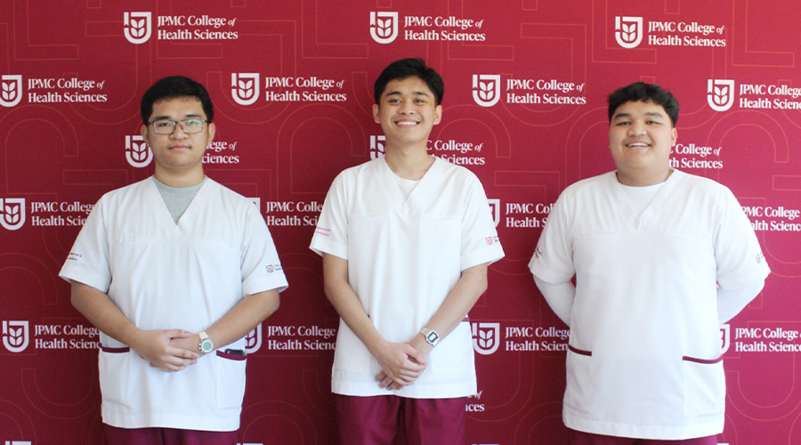 Nursing in Brunei | A group of three male nursing students wearing watch smiling to camera with JPMC College of Health Sciences backdrop in red and white