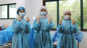 Nursing School in Asia | Three female nursing students wearing hospital gowns or scrubs and latex gloves doing the doctor pose in a classroom prior to starting clinical training